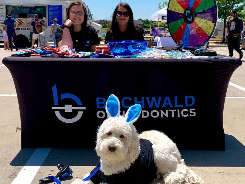 Fluffy white dog wearing blue bunny ears sitting by table for Buchwald Orthodontics at outdoor event